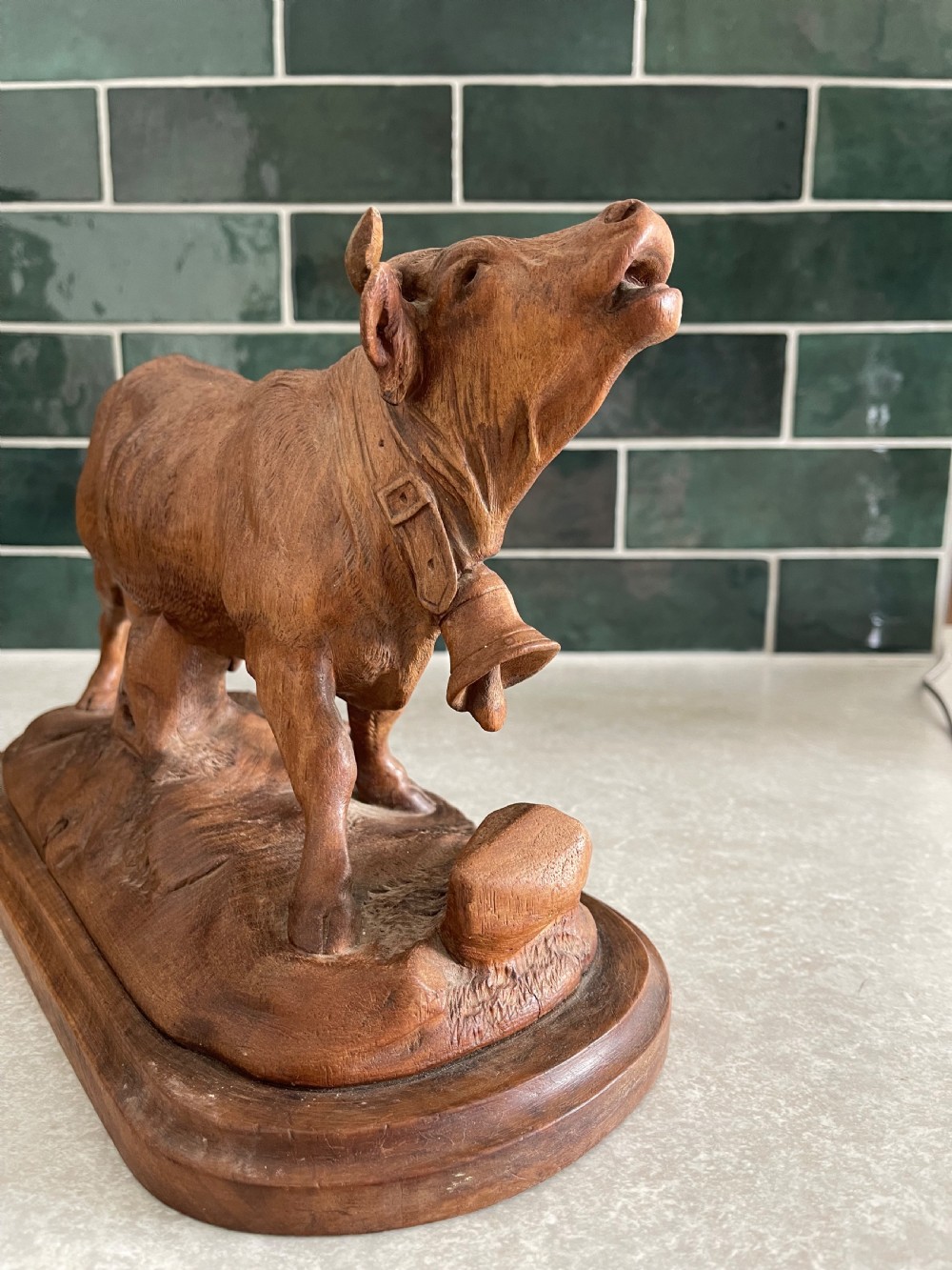 A detailed 19th-century carved wooden cow featuring an upturned head with a cowbell around its neck, standing on a carved naturalistic terrain base. View III