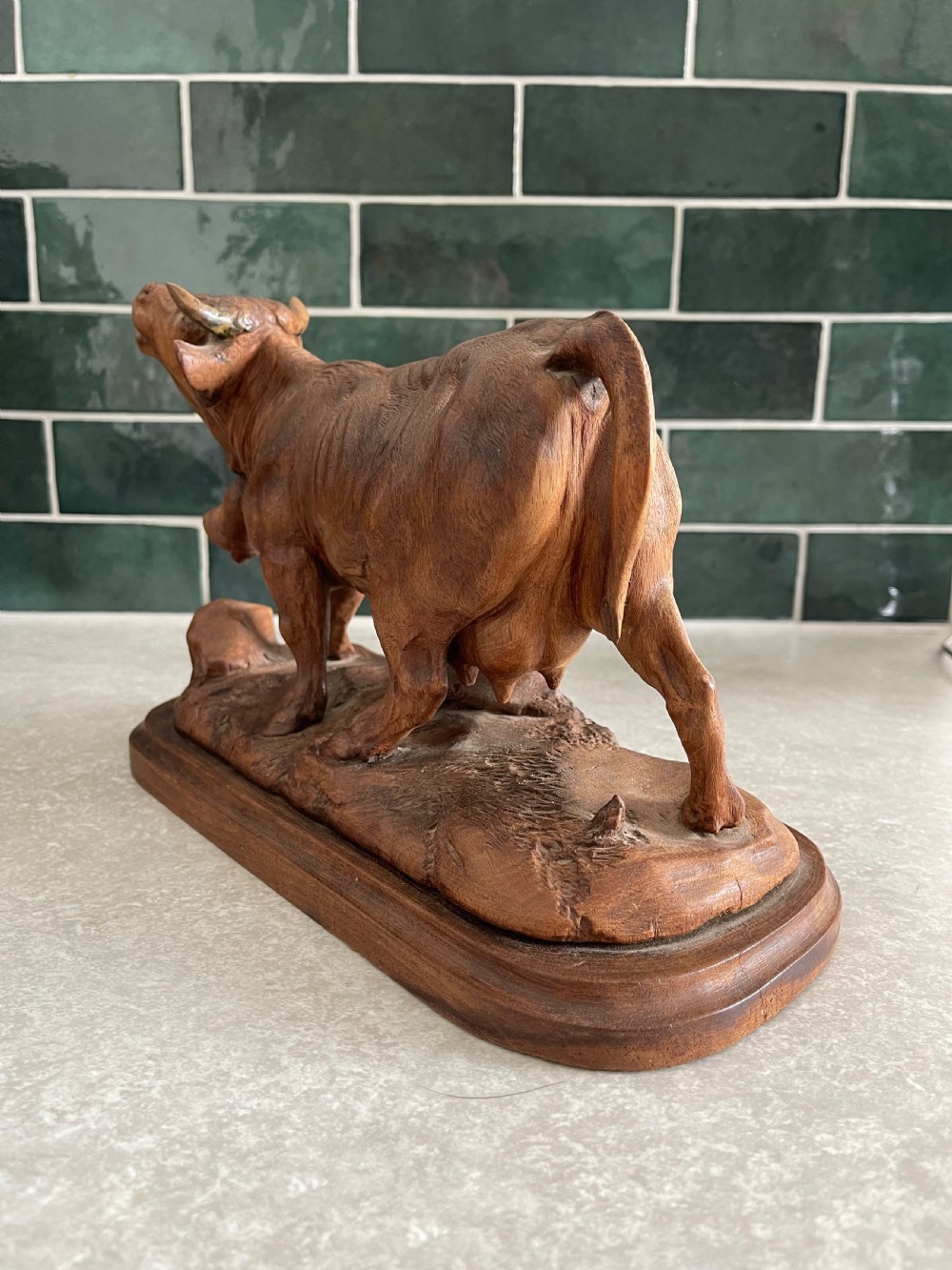 A detailed 19th-century carved wooden cow featuring an upturned head with a cowbell around its neck, standing on a carved naturalistic terrain base. View V