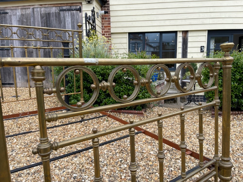 A Victorian single brass bed frame featuring a tall headboard and shorter footboard with turned finials and decorative circular ring inserts, set on castors. View II