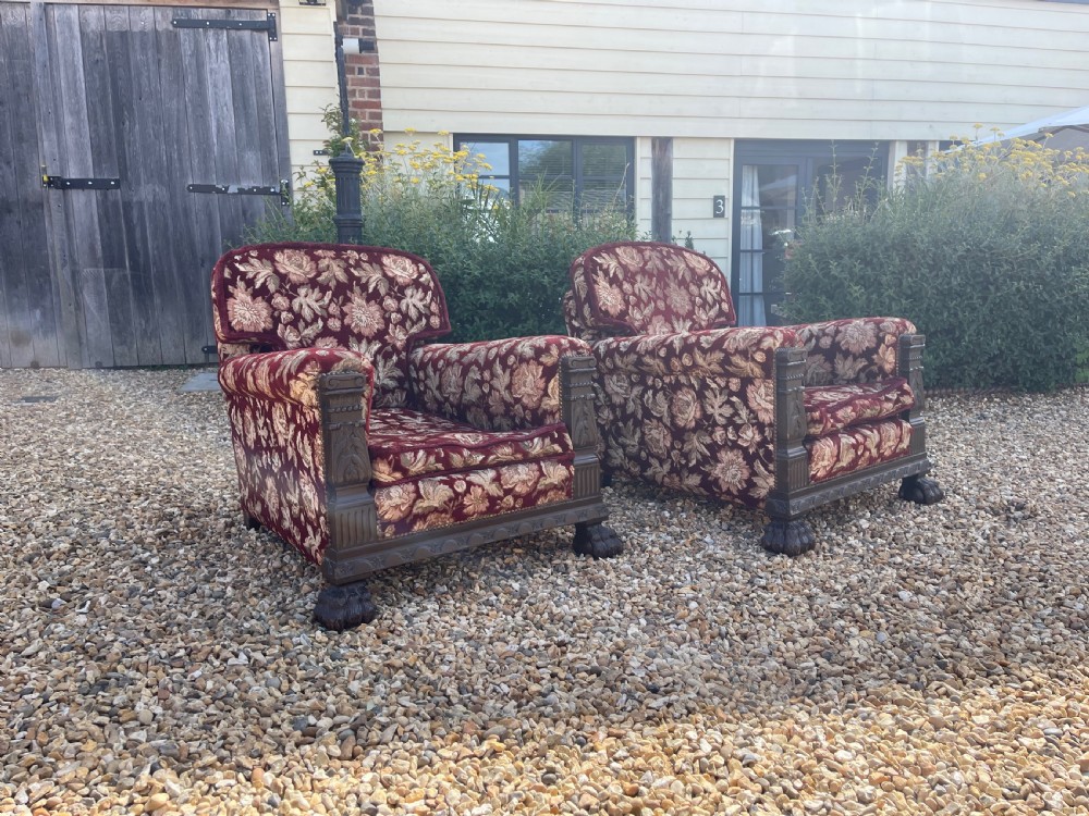 A pair of low-slung, deep-seated armchairs upholstered in burgundy and gold floral velvet with carved oak facings and claw feet.