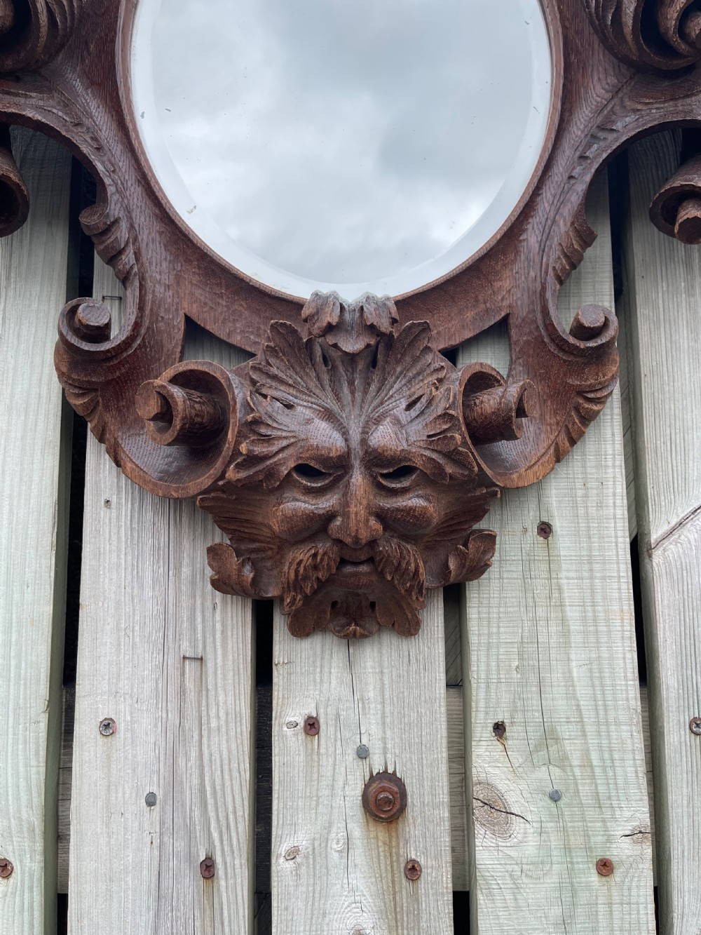 A nineteenth-century carved oak wall mirror featuring an oval glass plate framed by ornate scrollwork, acanthus leaves, and a carved face at the base. View III