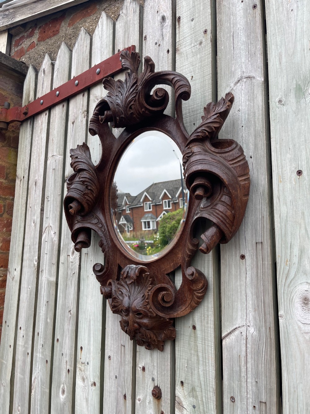 A nineteenth-century carved oak wall mirror featuring an oval glass plate framed by ornate scrollwork, acanthus leaves, and a carved face at the base. View V