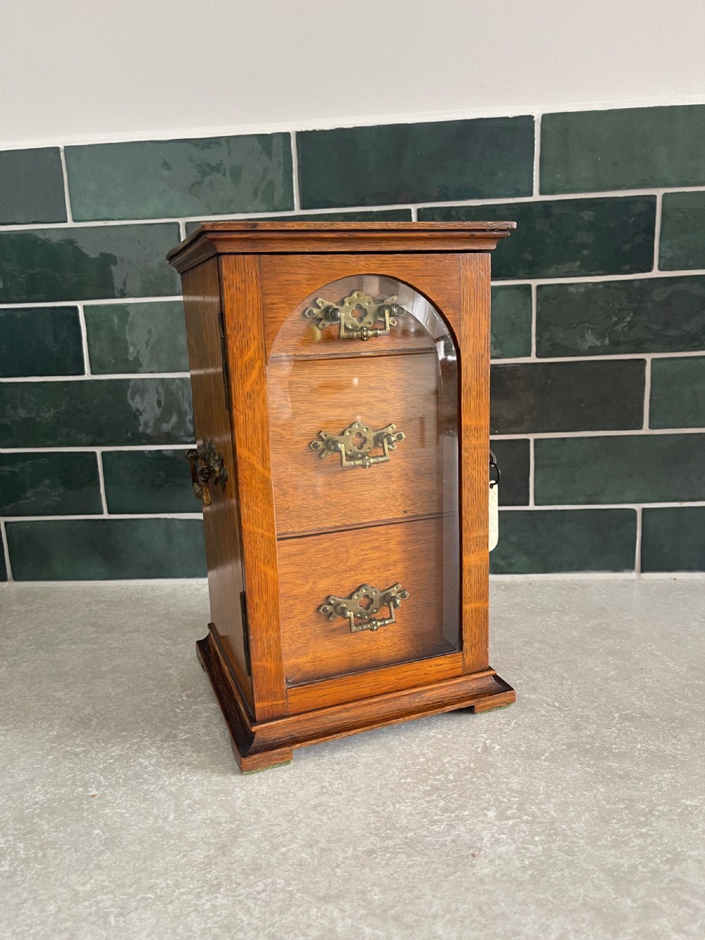 A 19th-century oak tabletop cabinet featuring three internal drawers behind an arched glazed door with decorative brass drop handles.