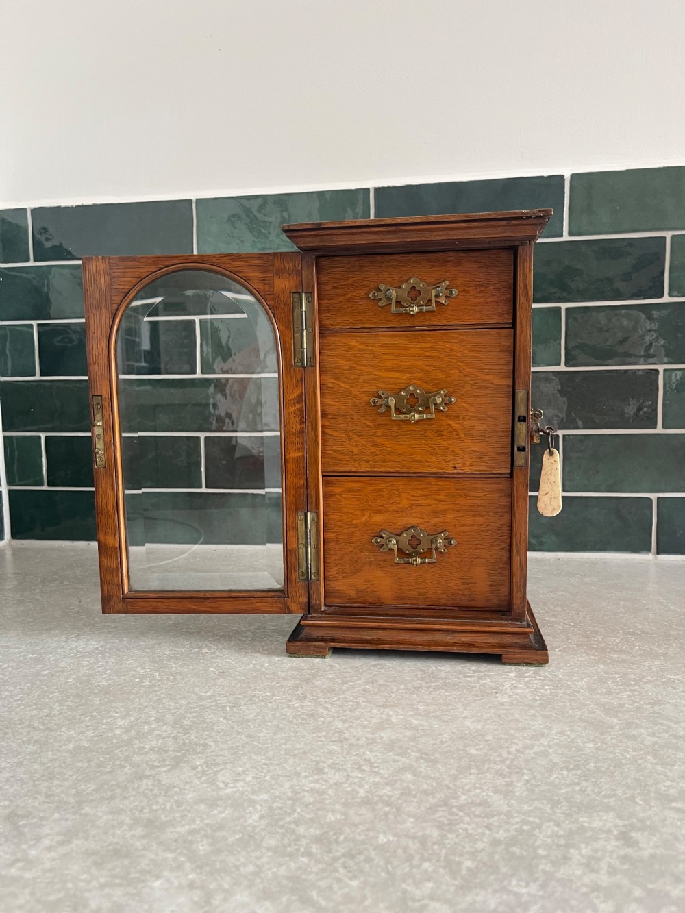 A 19th-century oak tabletop cabinet featuring three internal drawers behind an arched glazed door with decorative brass drop handles. View II