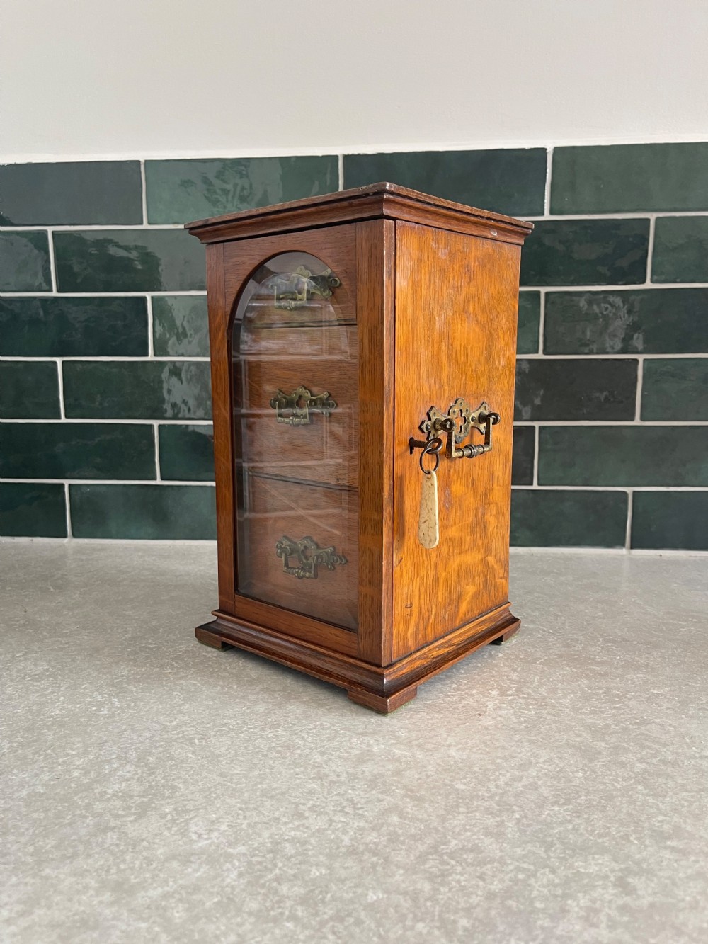 A 19th-century oak tabletop cabinet featuring three internal drawers behind an arched glazed door with decorative brass drop handles. View III
