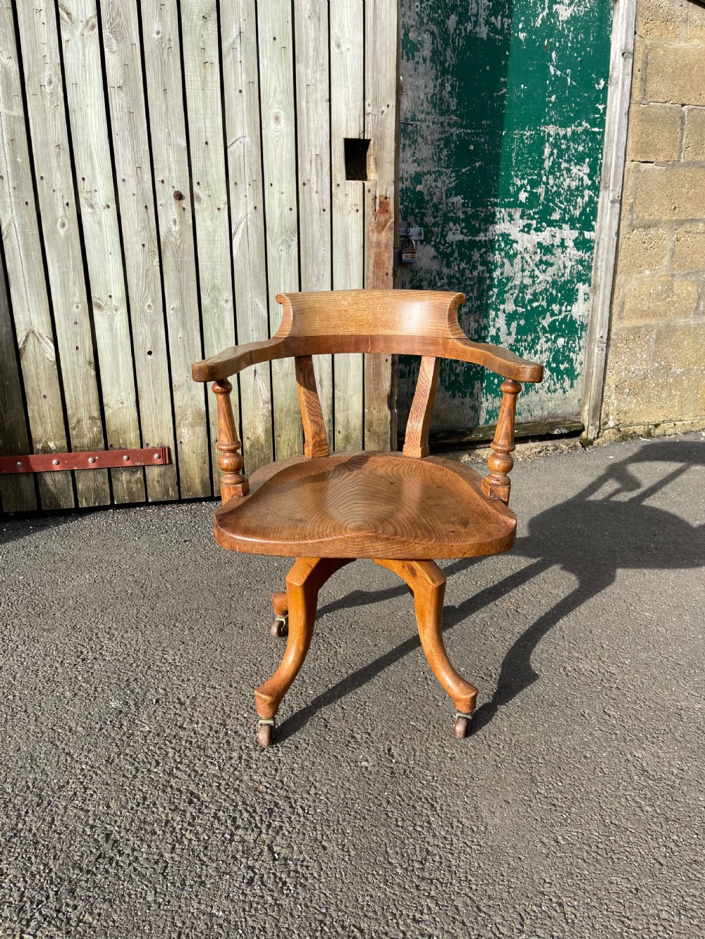 An antique golden oak revolving desk chair featuring curved armrests, a contoured wooden seat, and a four-legged base on castors.