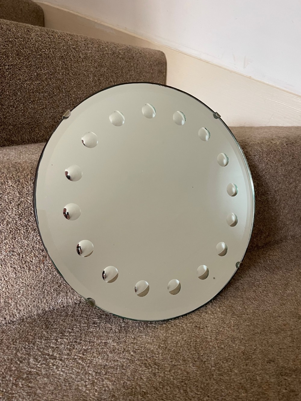 A circular frameless glass wall mirror featuring a ring of carved concave roundels around its perimeter.
