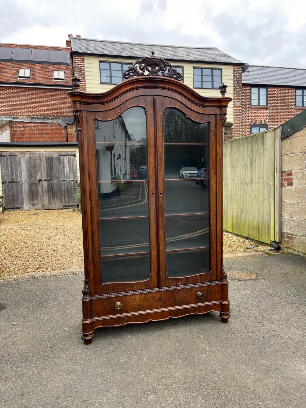 A tall dark rosewood Continental display cabinet featuring arched glazed double doors, an intricate carved pediment cresting, and a lower drawer.