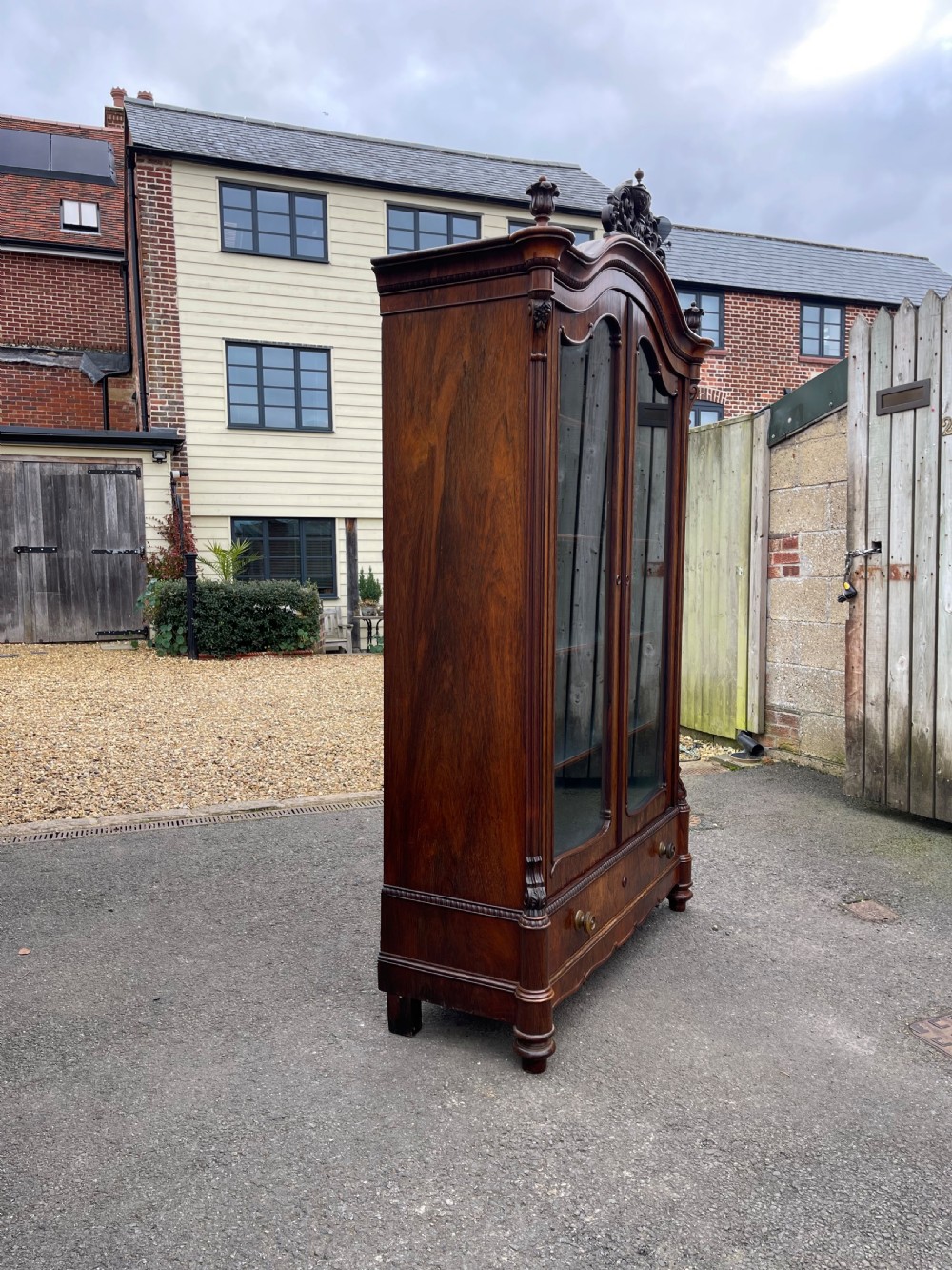 A tall dark rosewood Continental display cabinet featuring arched glazed double doors, an intricate carved pediment cresting, and a lower drawer. View VI