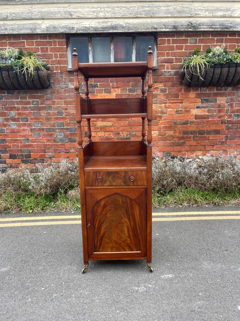 A nineteenth-century mahogany cupboard base whatnot featuring open shelves supported by turned posts, a single drawer, and a lower cabinet with an arched flame mahogany panel on brass castors.