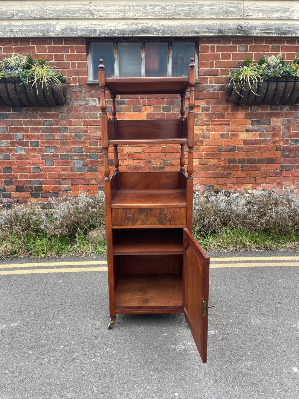 A nineteenth-century mahogany cupboard base whatnot featuring open shelves supported by turned posts, a single drawer, and a lower cabinet with an arched flame mahogany panel on brass castors. View IV