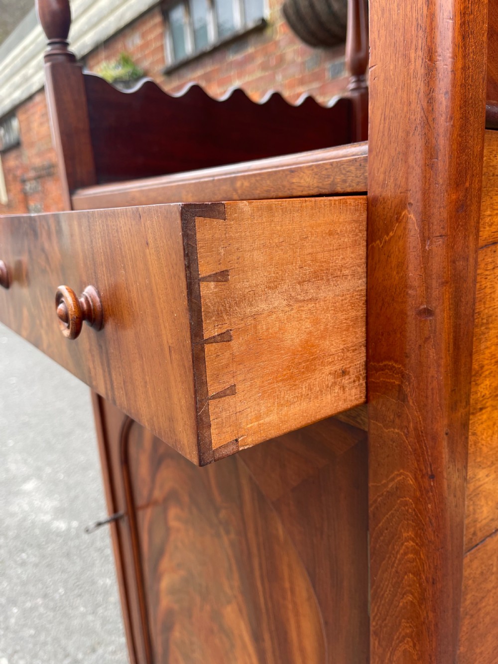 A nineteenth-century mahogany cupboard base whatnot featuring open shelves supported by turned posts, a single drawer, and a lower cabinet with an arched flame mahogany panel on brass castors. View V
