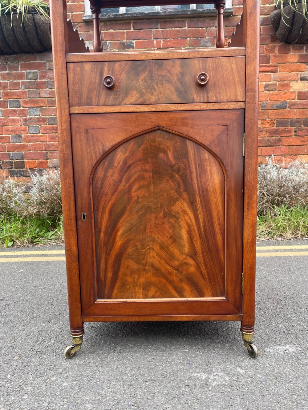 A nineteenth-century mahogany cupboard base whatnot featuring open shelves supported by turned posts, a single drawer, and a lower cabinet with an arched flame mahogany panel on brass castors. View IX
