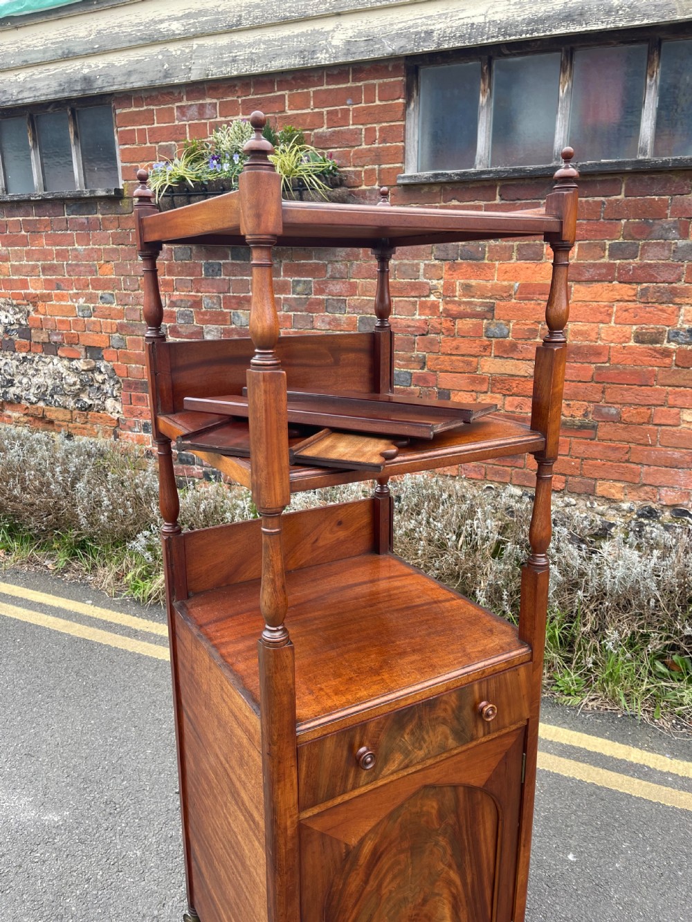 A nineteenth-century mahogany cupboard base whatnot featuring open shelves supported by turned posts, a single drawer, and a lower cabinet with an arched flame mahogany panel on brass castors. View X