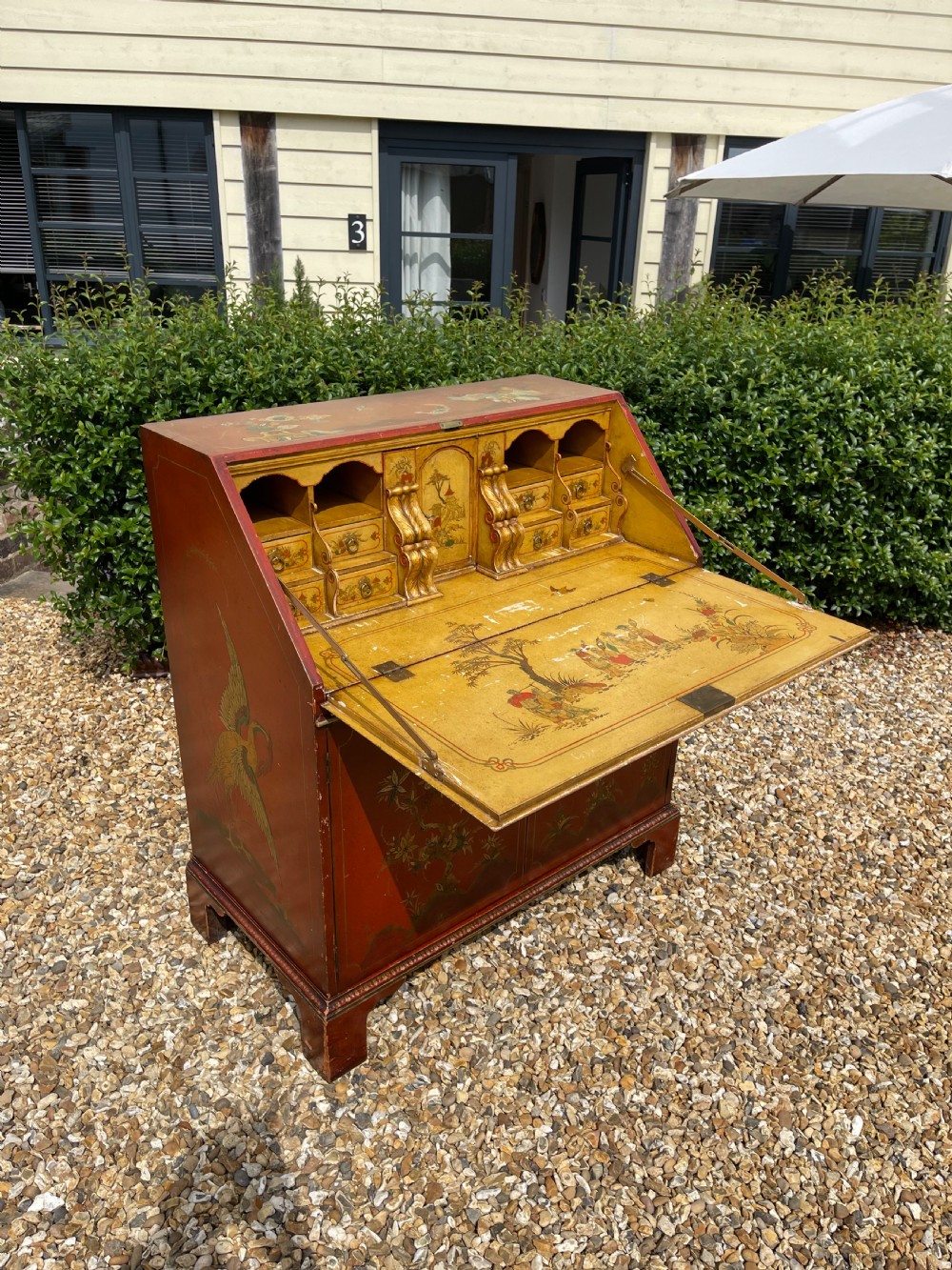 An antique red lacquered fall-front bureau with hand-painted Chinoiserie scenes and a yellow fitted interior featuring cubbies and small drawers.