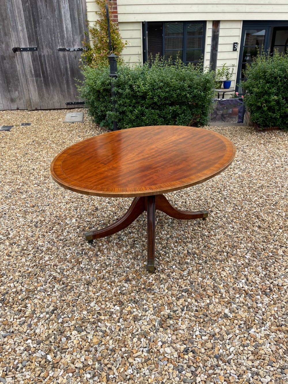An oval George III mahogany breakfast table with crossbanded satinwood border on a central pedestal base with four splayed legs terminating in brass castors.