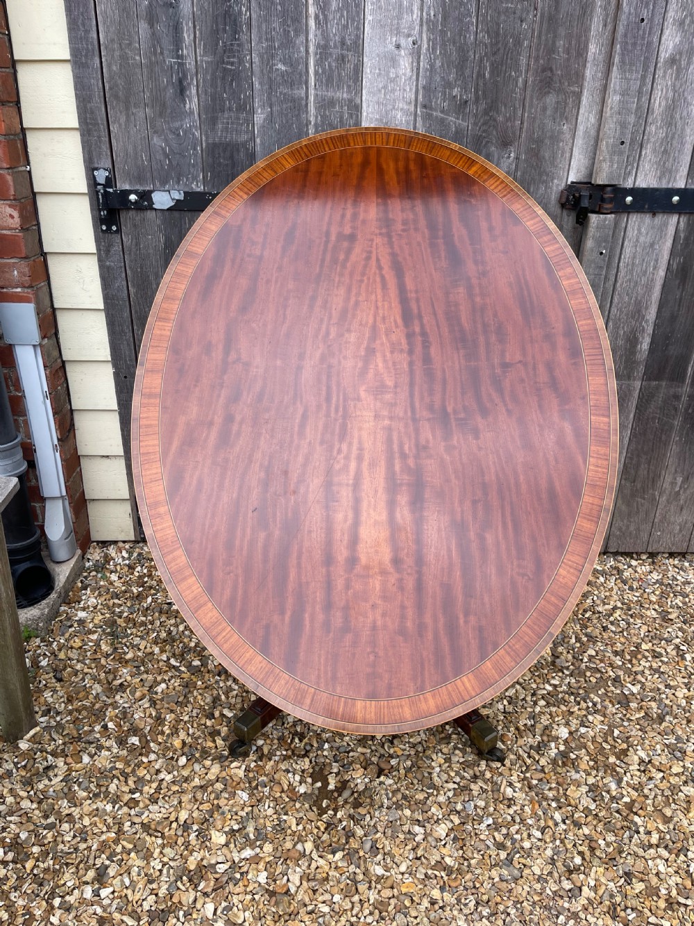 An oval George III mahogany breakfast table with crossbanded satinwood border on a central pedestal base with four splayed legs terminating in brass castors. View III
