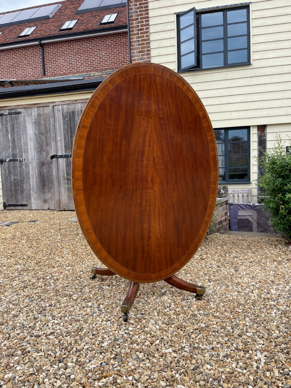An oval George III mahogany breakfast table with crossbanded satinwood border on a central pedestal base with four splayed legs terminating in brass castors. View V