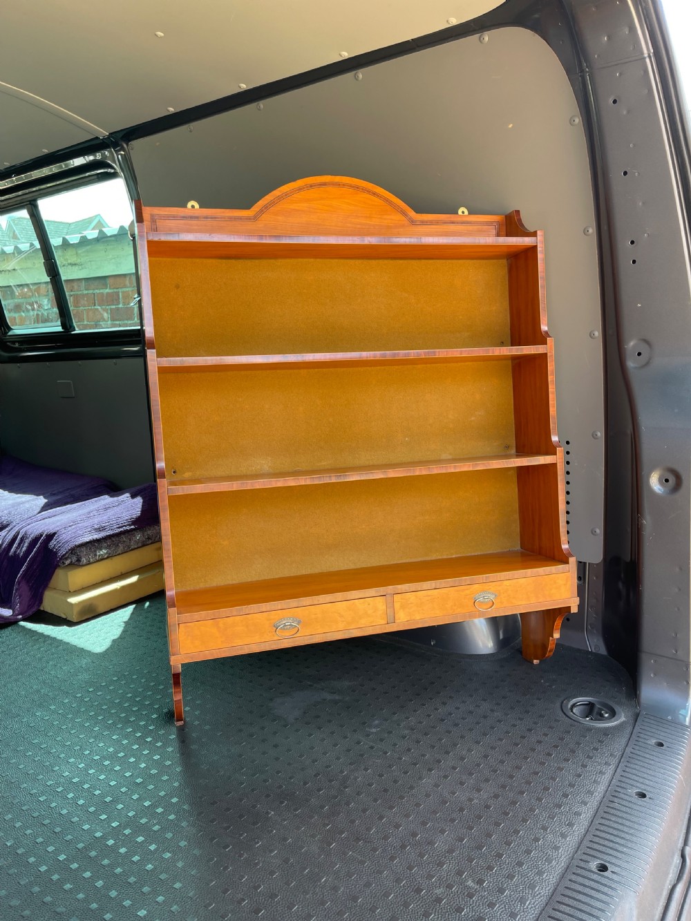 An antique satinwood hanging shelf unit featuring an arched cresting rail, three open tiers, and two drawers at the base with brass drop ring handles.