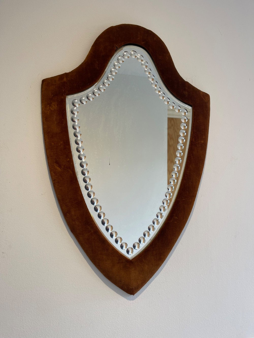 A shield-shaped wall mirror featuring a warm brown velvet fabric frame and an inner border of decorative circular glass beads.