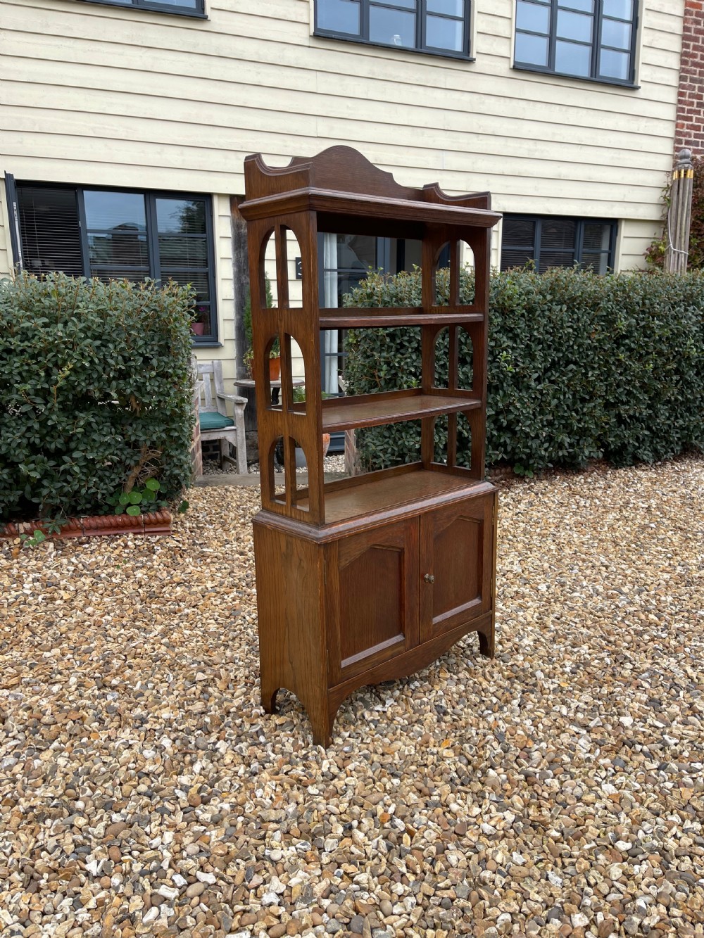 A freestanding oak open bookcase featuring two upper shelves with arched side cutouts over a lower two-door cupboard.