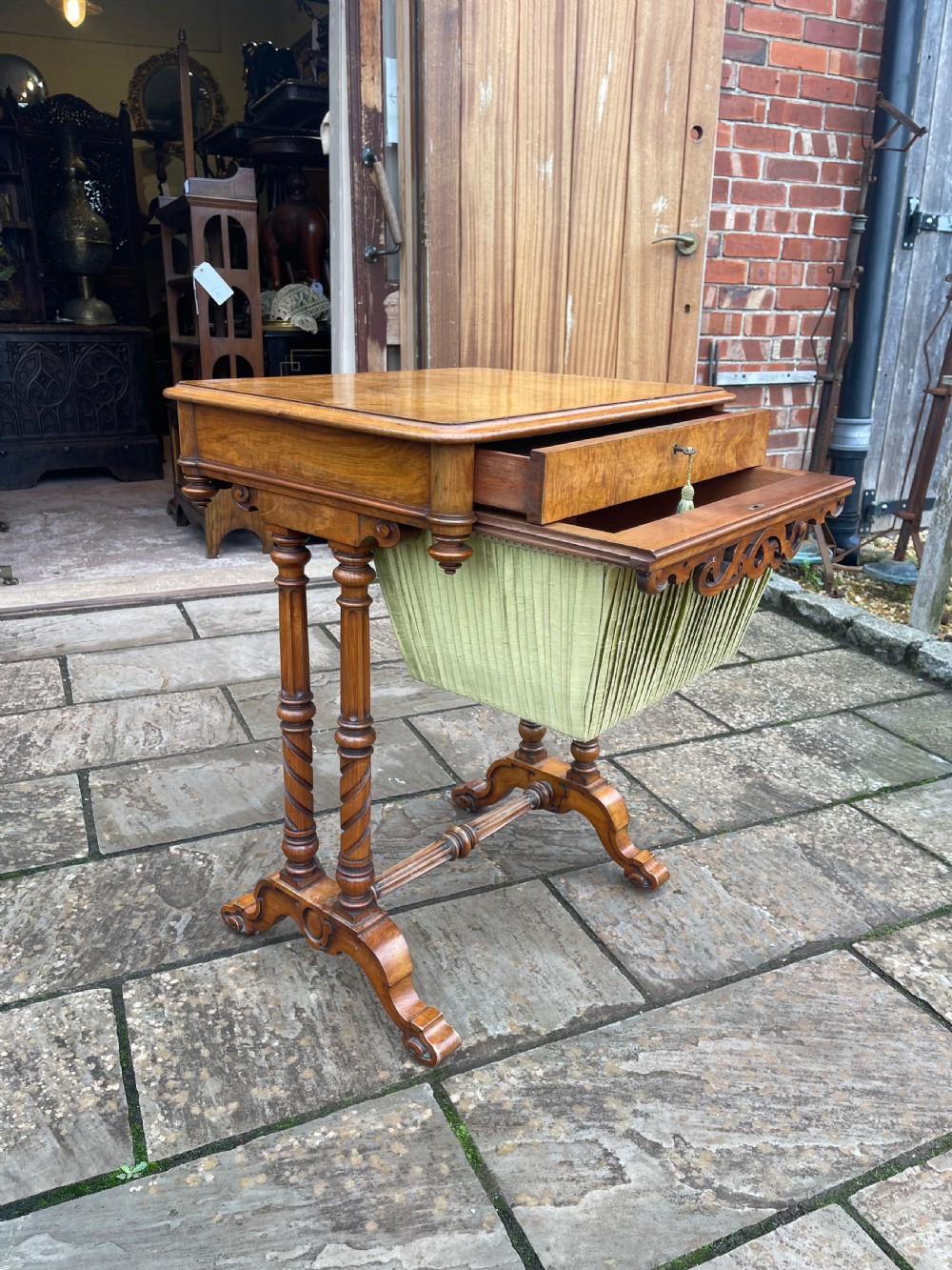A Victorian burr walnut sewing work table with turned barley twist columns, a pleated green fabric basket, and a carved trestle base. View VI
