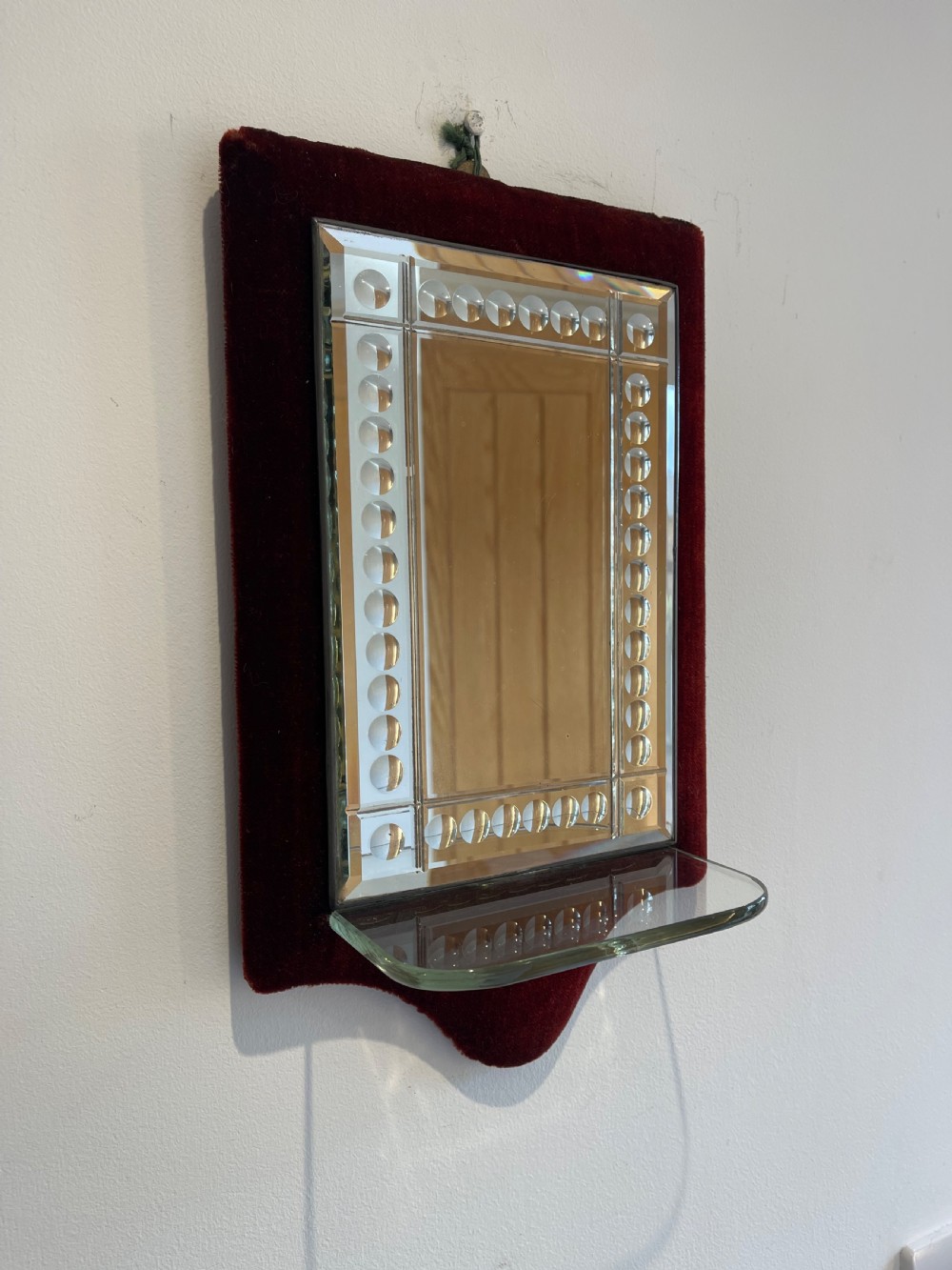 A rectangular bevelled glass mirror with cut roundel details, mounted on a deep red velvet backing panel with an attached clear glass shelf.