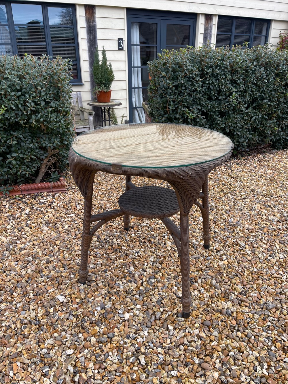 A round dark brown Lloyd Loom table featuring a circular glass top protective surface, flared woven legs, and a lower central woven shelf.