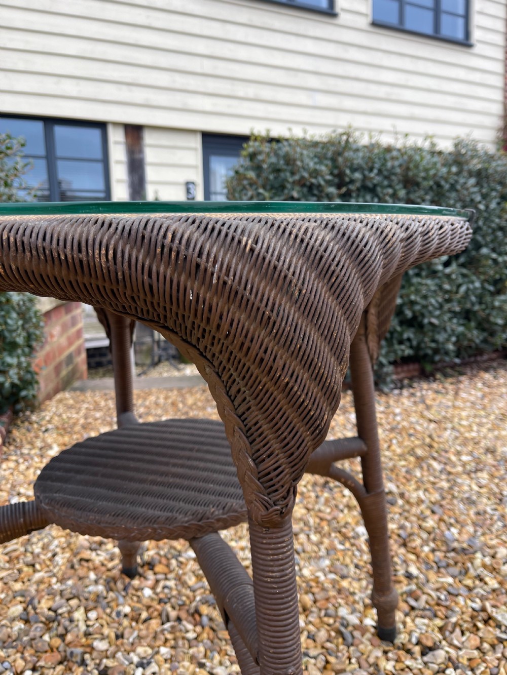 A round dark brown Lloyd Loom table featuring a circular glass top protective surface, flared woven legs, and a lower central woven shelf. View VI
