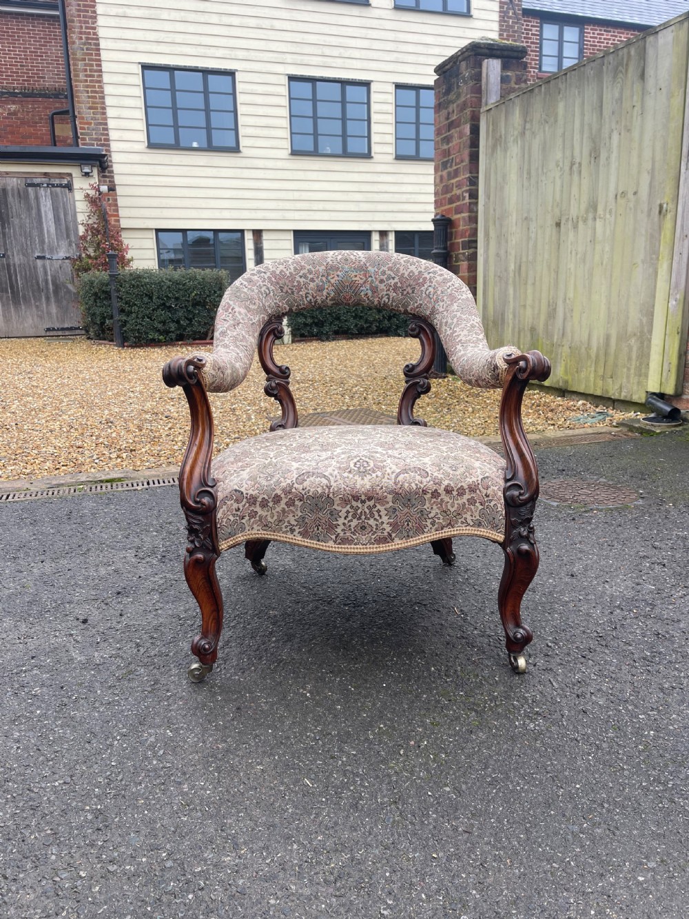 An early Victorian tub armchair crafted from dark carved rosewood with scroll armrests, cabriole legs on brass castors, and upholstered in a patterned beige floral fabric.