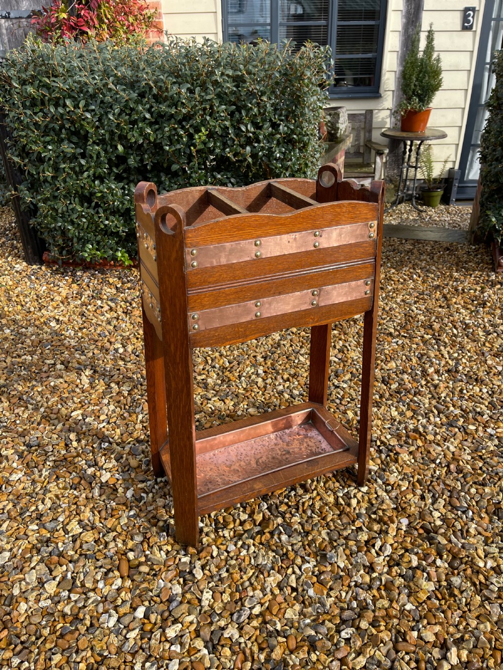 An oak Arts and Crafts umbrella stand featuring hammered copper bands, circular pierced side handles, four top compartments, and a removable copper drip tray at the base.