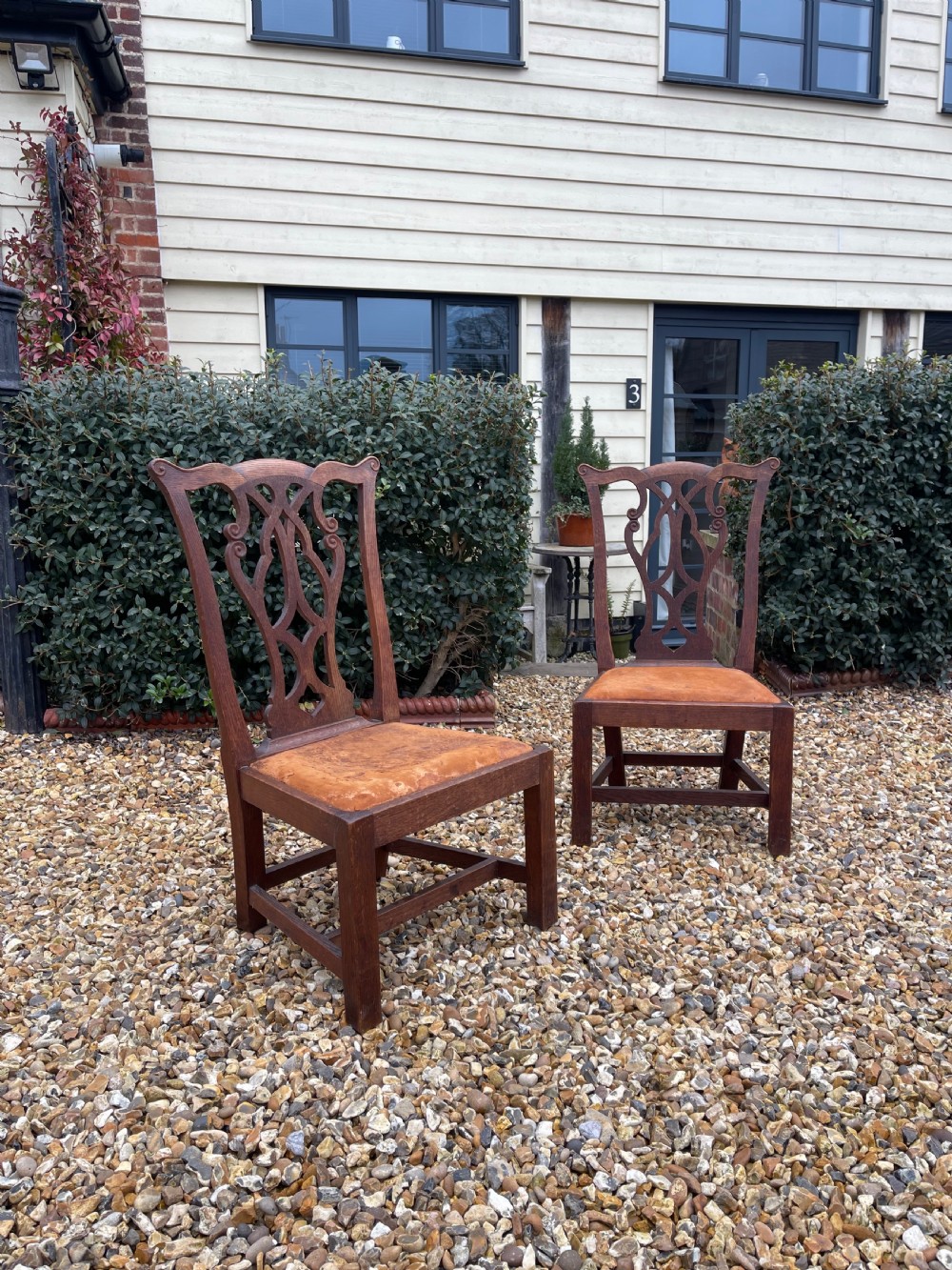 A pair of dark oak antique side chairs with intricately carved openwork back splats and tan upholstered seats.