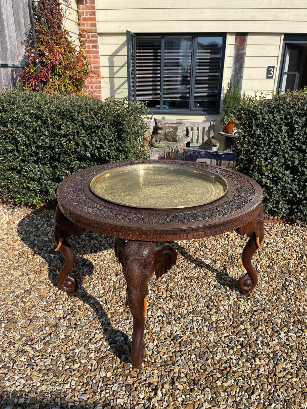 A circular carved dark wood table featuring legs shaped as elephant heads with tusks and an inset circular engraved brass tray in the centre.