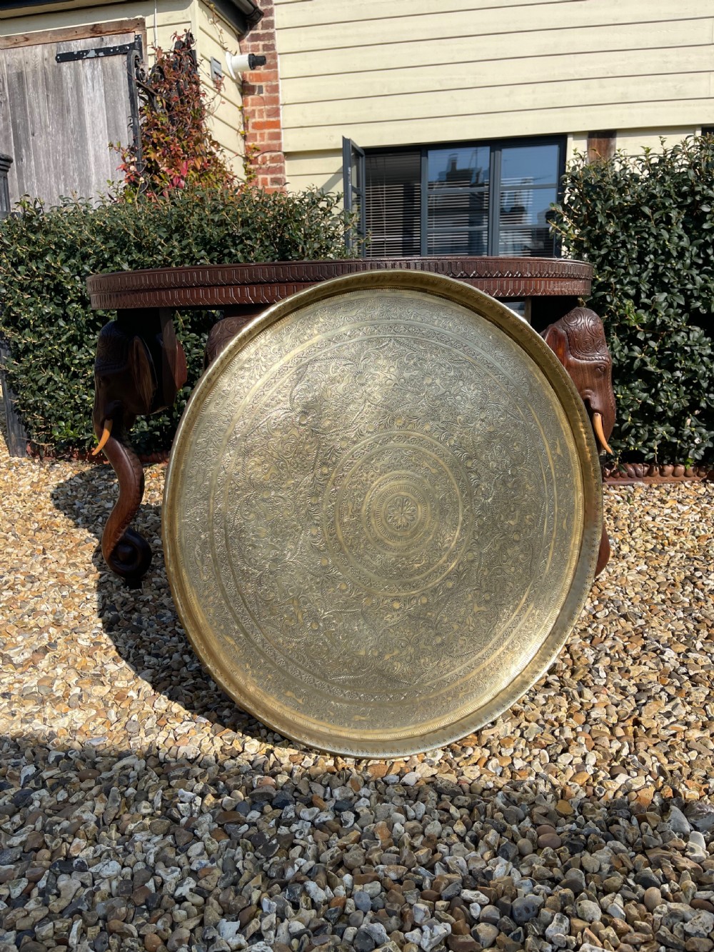 A circular carved dark wood table featuring legs shaped as elephant heads with tusks and an inset circular engraved brass tray in the centre. View IV