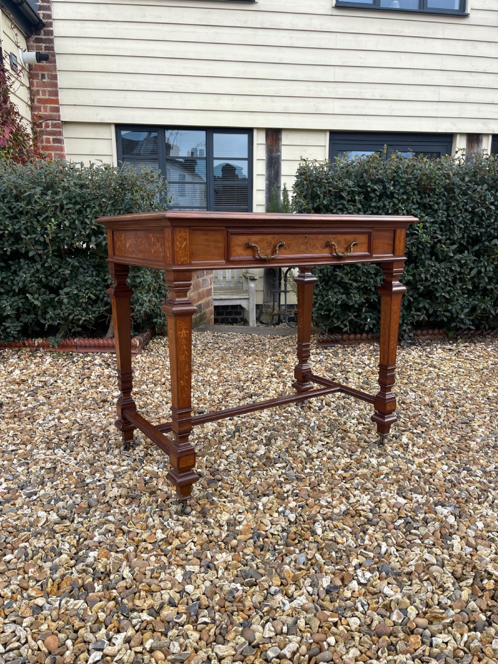 A Victorian inlaid mahogany writing table featuring a single drawer with brass drop handles, floral marquetry, square tapered legs, and an H-stretcher base.
