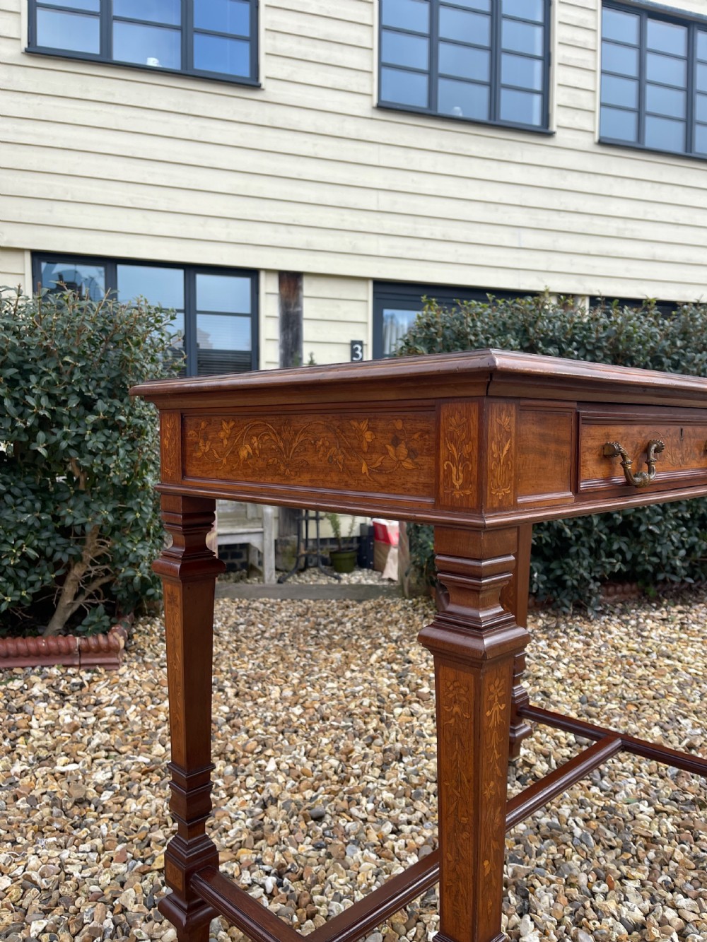A Victorian inlaid mahogany writing table featuring a single drawer with brass drop handles, floral marquetry, square tapered legs, and an H-stretcher base. View VI