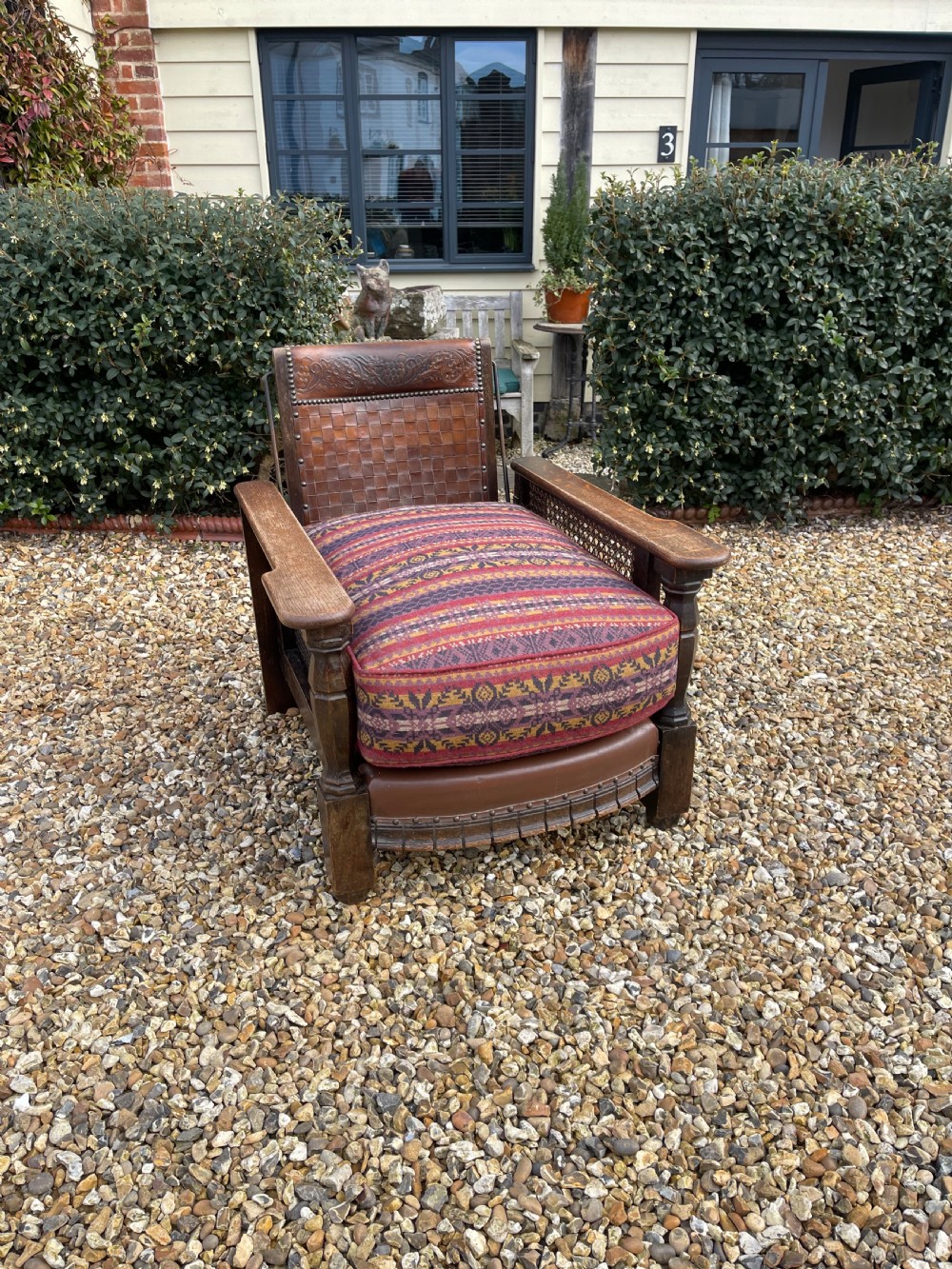 An antique Arts and Crafts oak reclining armchair featuring wide flat armrests, a woven leather back, caned sides, and a pattern-upholstered cushion.