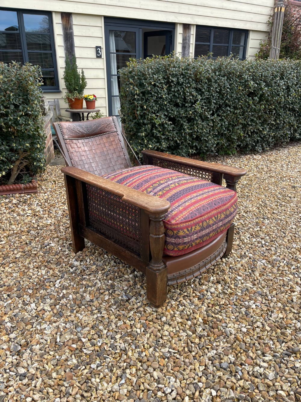 An antique Arts and Crafts oak reclining armchair featuring wide flat armrests, a woven leather back, caned sides, and a pattern-upholstered cushion. View V