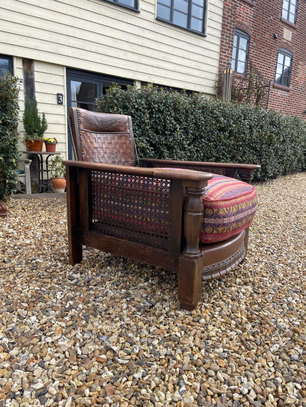 An antique Arts and Crafts oak reclining armchair featuring wide flat armrests, a woven leather back, caned sides, and a pattern-upholstered cushion. View VI