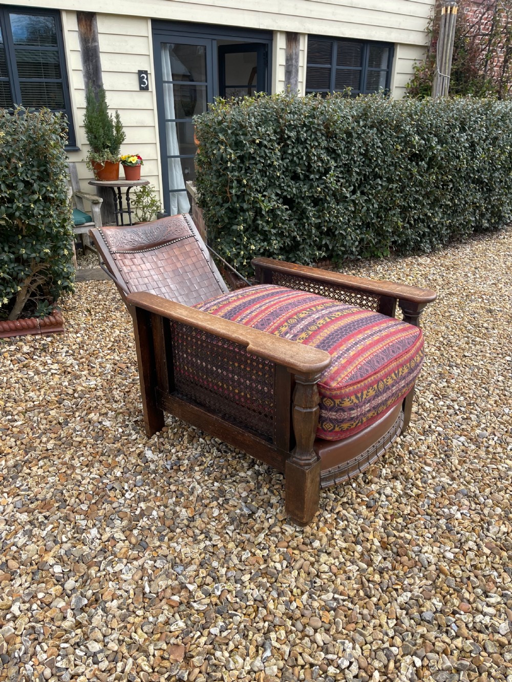 An antique Arts and Crafts oak reclining armchair featuring wide flat armrests, a woven leather back, caned sides, and a pattern-upholstered cushion. View VII