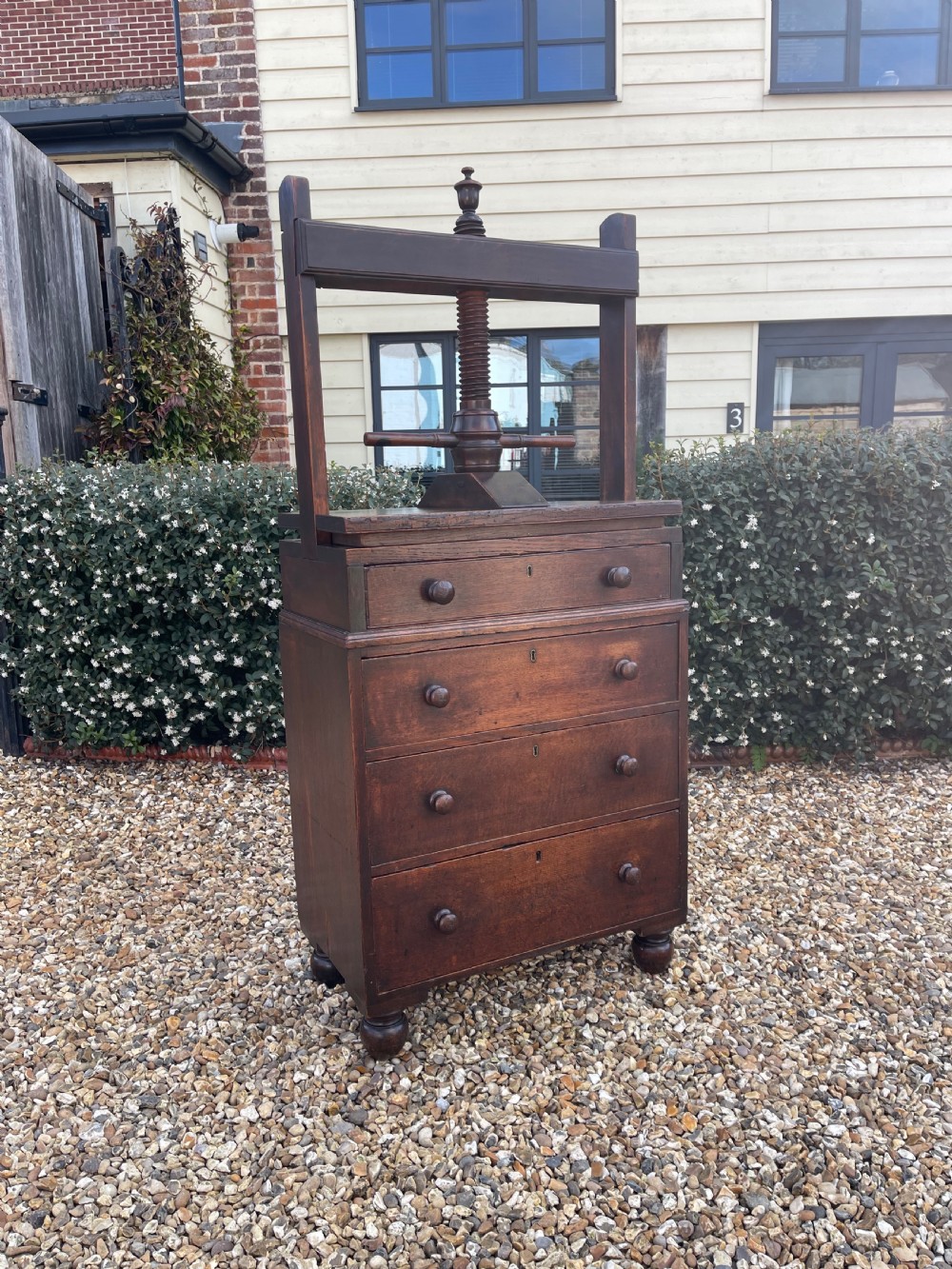 A dark oak press chest with four graduated drawers, turned knobs, bun feet, and a top-mounted wooden screw mechanism.