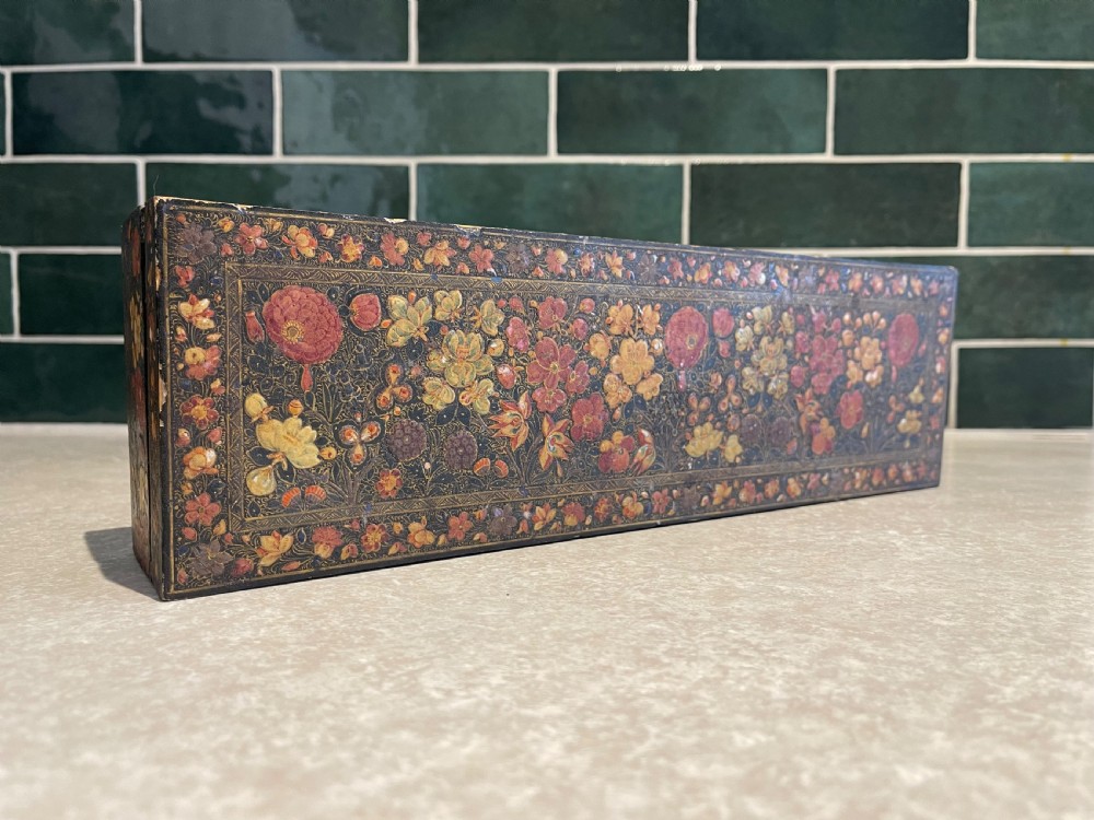 A long rectangular papier-mache box with intricate hand-painted floral patterns in red, yellow, and gold lacquer over a dark background.