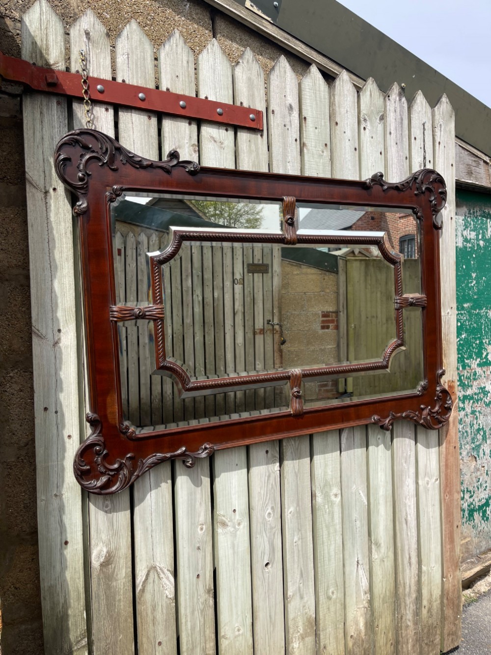 A large horizontal rectangular wall mirror framed in polished dark mahogany with ornate carved scrollwork corners and a central beaded border surrounding multi-panel sectional glass.