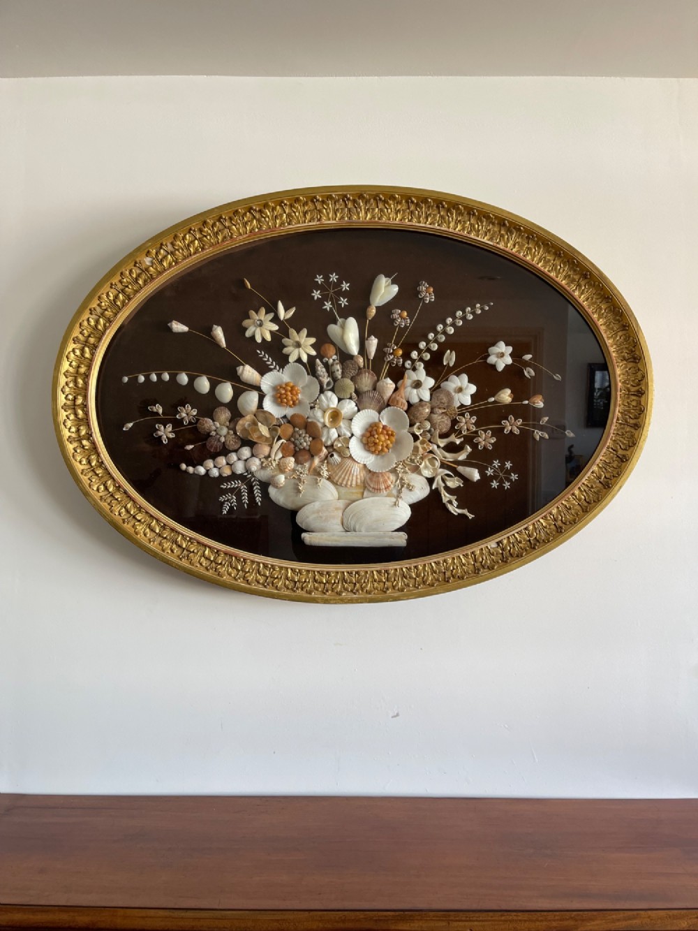 An oval gilt-framed picture featuring a three-dimensional bouquet of sea shells arranged into flowers against a dark brown background.