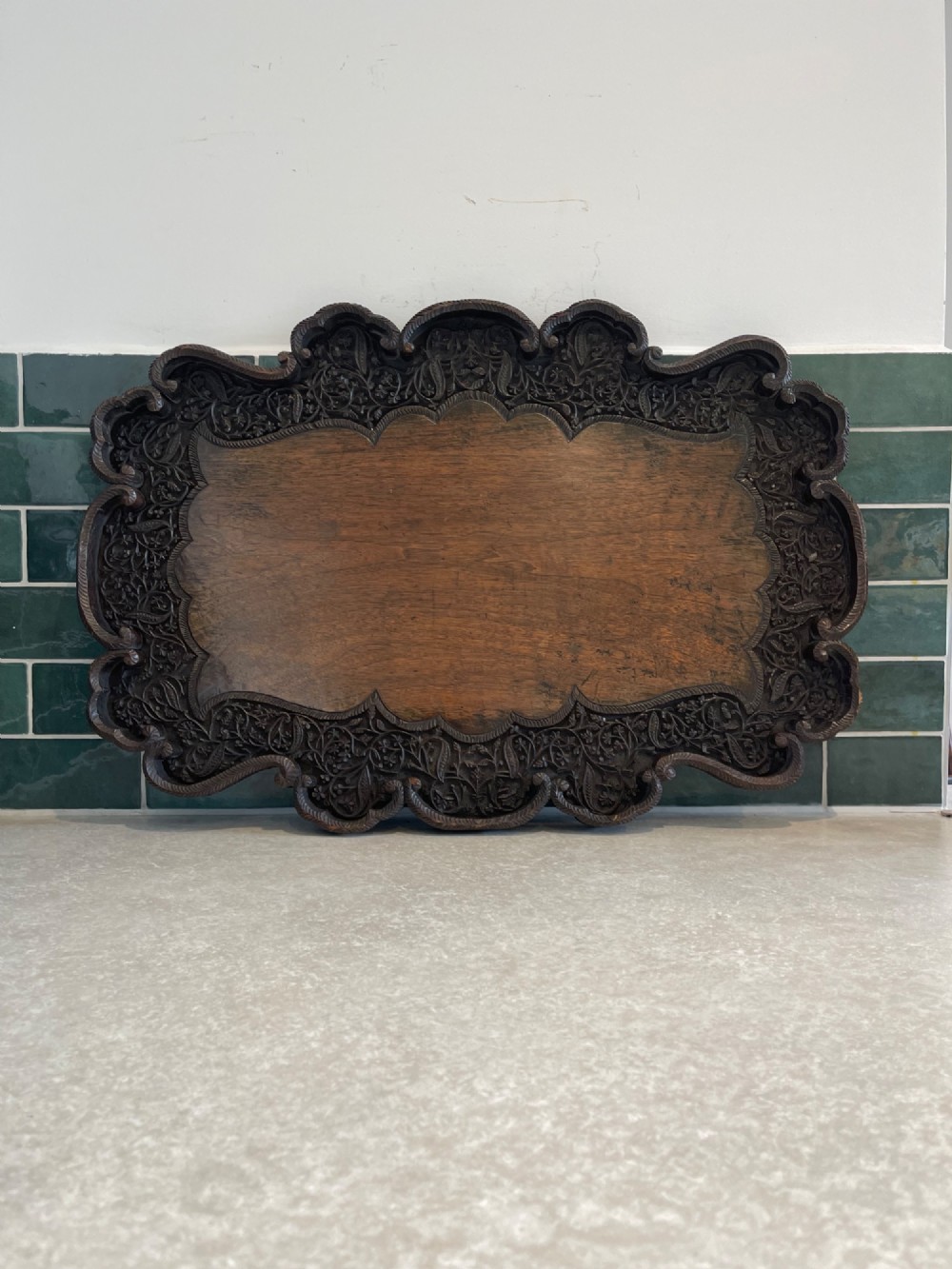 A dark brown carved walnut tray featuring a smooth central panel surrounded by a scalloped, intricately carved floral border.