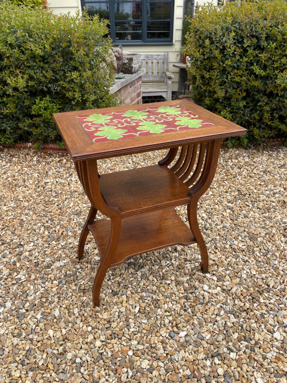 An antique oak side table featuring an inset top panel of six green and terracotta glazed tiles with botanical motifs, raised on curved slatted supports and two lower shelves.