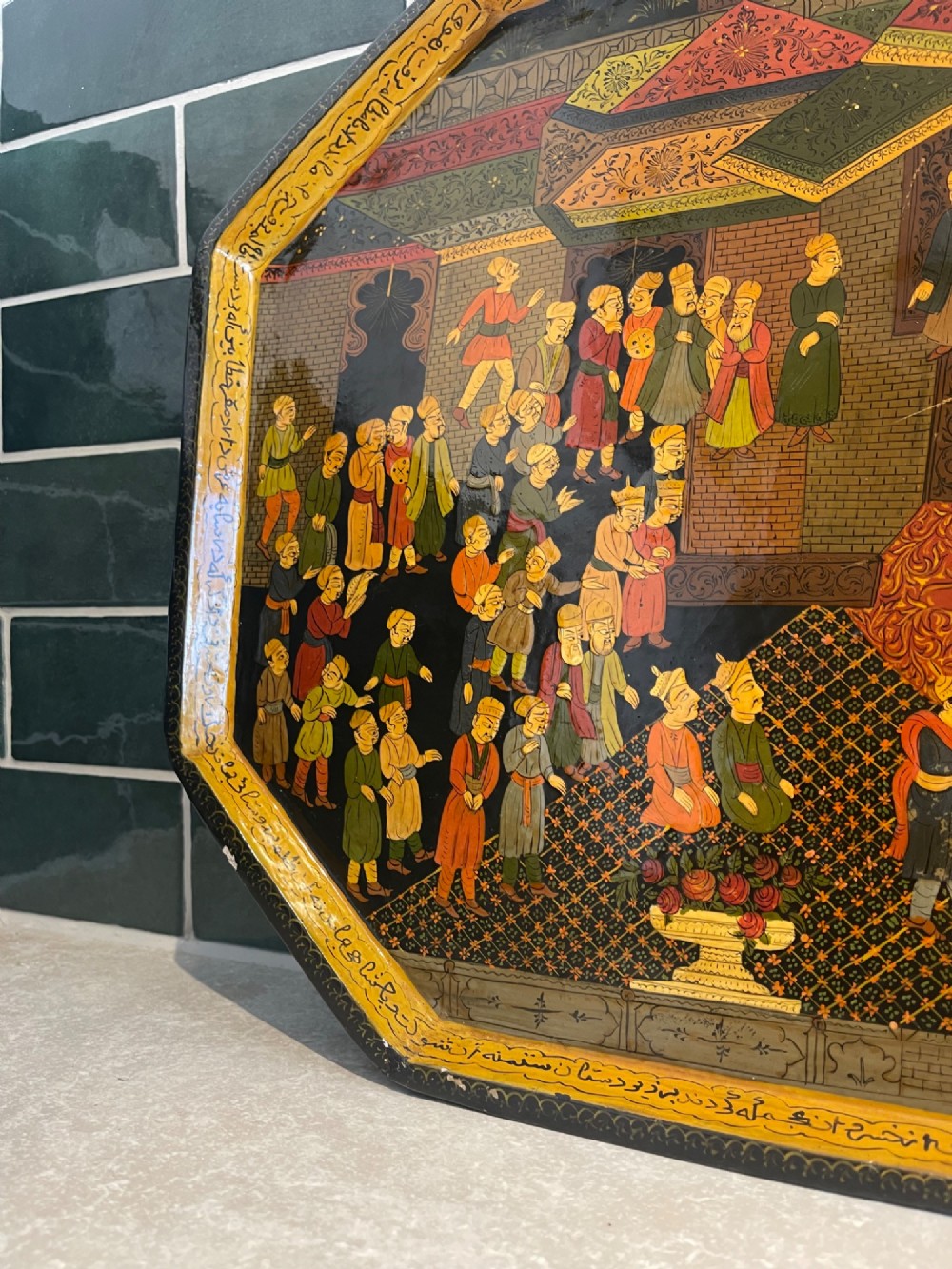 An octagonal Kashmiri papier-mache tray featuring a detailed hand-painted court scene with figures surrounded by a calligraphic border. View V