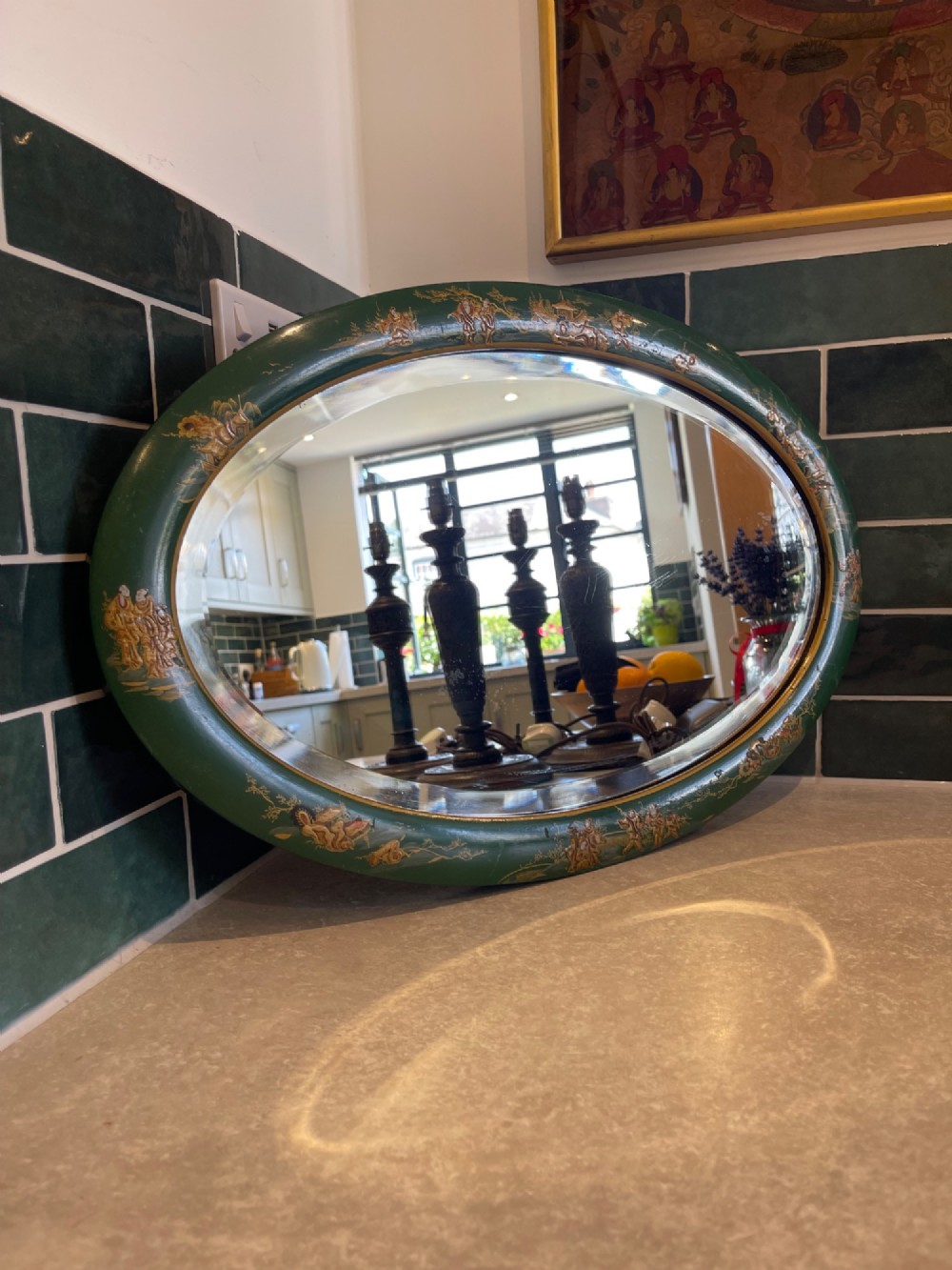 An oval wall mirror featuring a deep green lacquered wooden frame adorned with hand-painted gold chinoiserie figural landscapes.