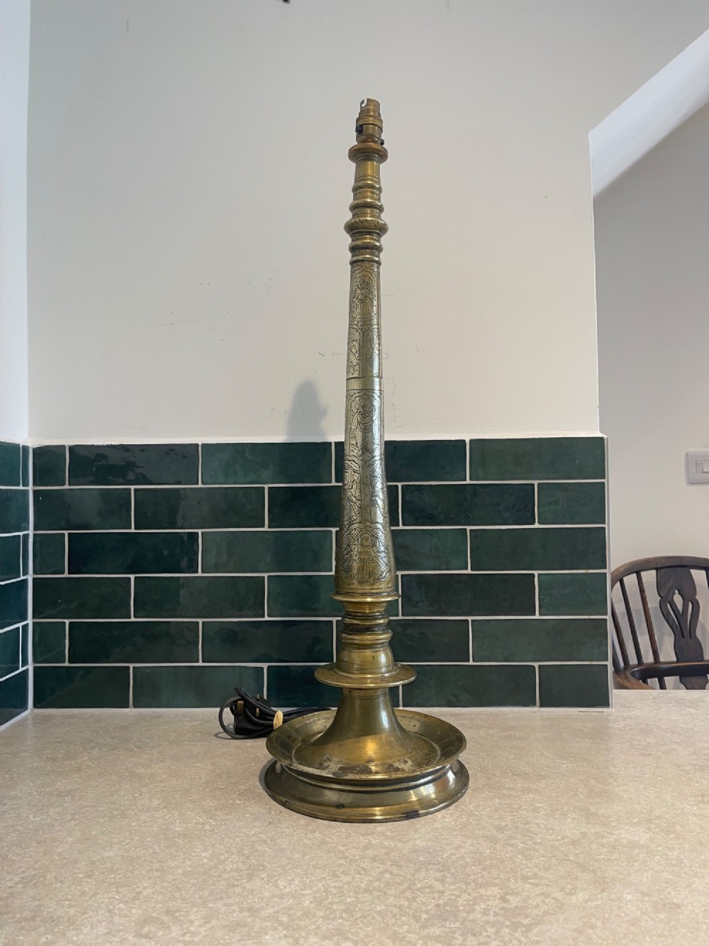 A tall, hand-engraved brass table lamp base featuring a tapered column, intricate etched figurative motifs, turned mouldings, and a stepped circular foot.