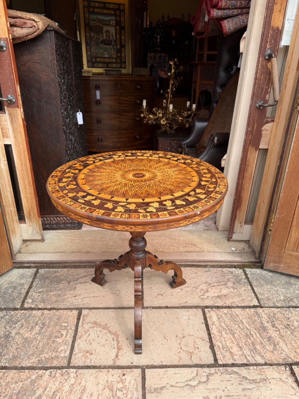 A circular wooden pedestal table featuring elaborate geometric starburst and floral vine marquetry inlays supported by a turned central column and carved tripod legs.